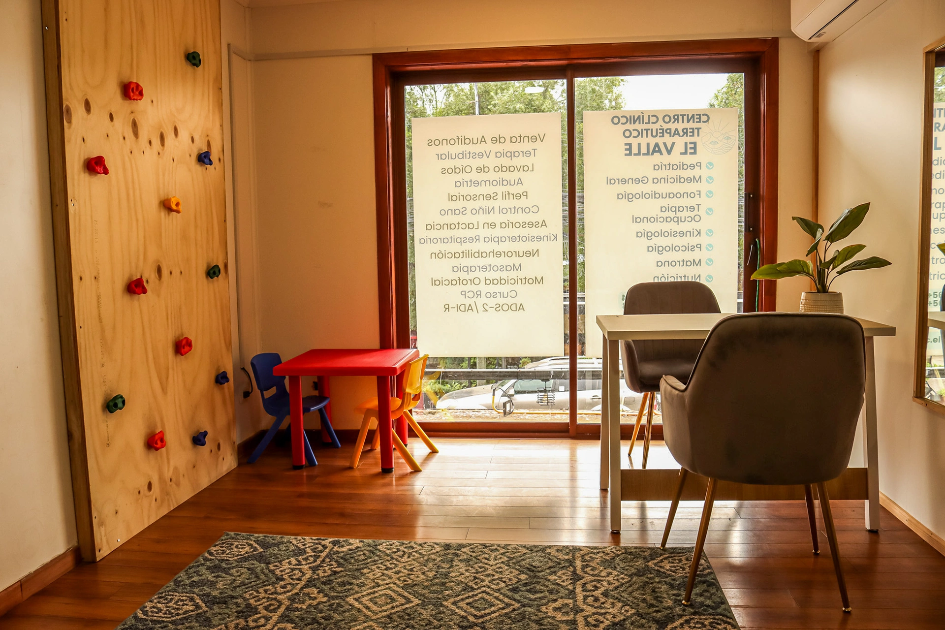 Sala de terapia con pared de escalada de madera, ventanal grande y zona de estar con mesa roja