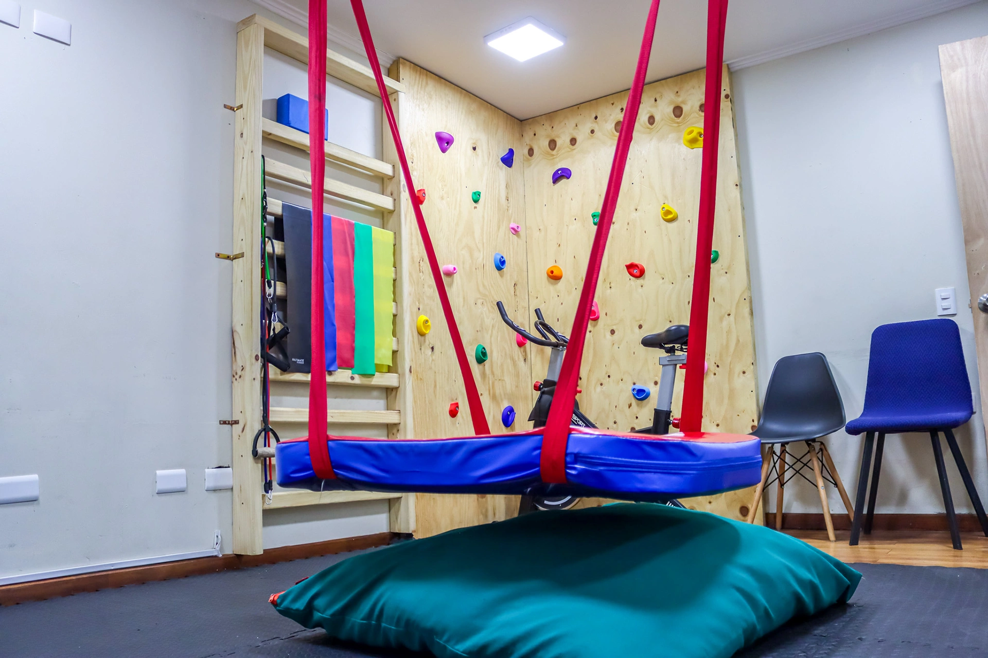 Sala de integración sensorial con hamaca azul colgada, pared de escalada al fondo y cojín en el piso
