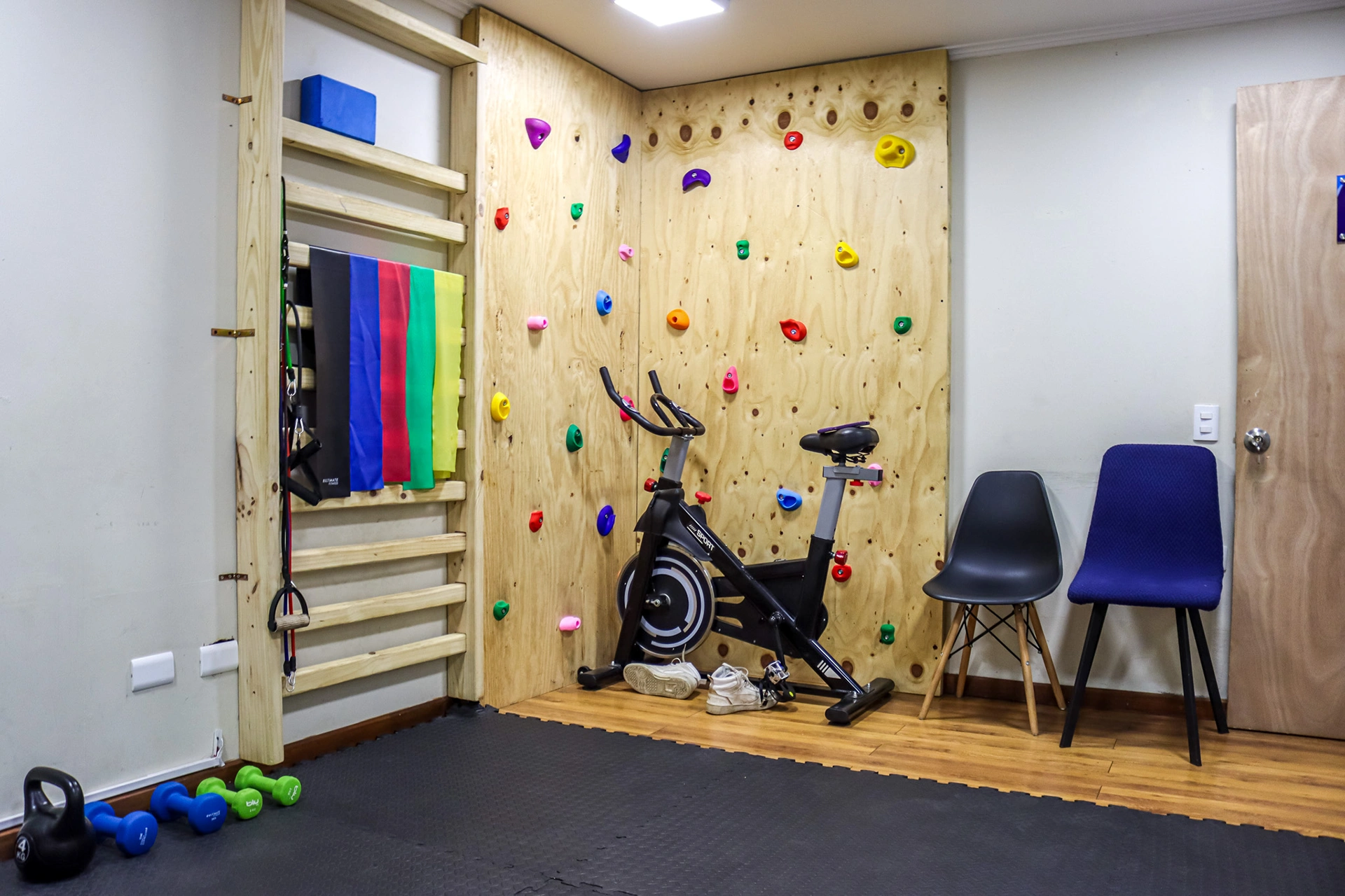 Sala de integración sensorial con pared de escalada con presas de colores, bicicleta estática y silla azul